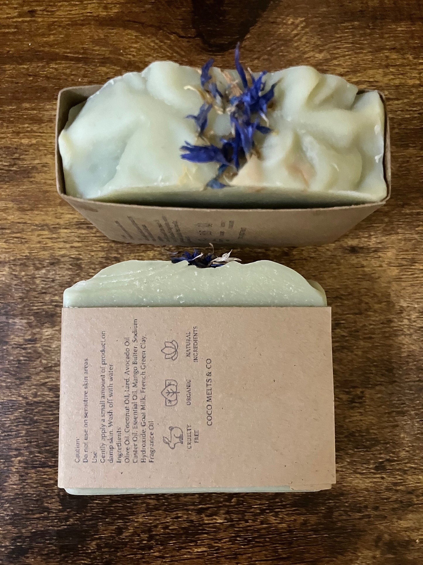 Handcrafted White Tea and Sage bar soap by Coco Melts and Co, topped with dried blue cornflowers, wrapped in kraft paper label, displayed on a cozy textured blanket with green leaves