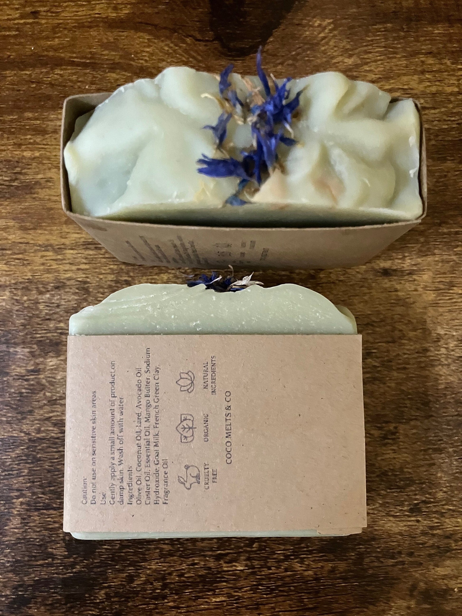 Handcrafted White Tea and Sage bar soap by Coco Melts and Co, topped with dried blue cornflowers, wrapped in kraft paper label, displayed on a cozy textured blanket with green leaves
